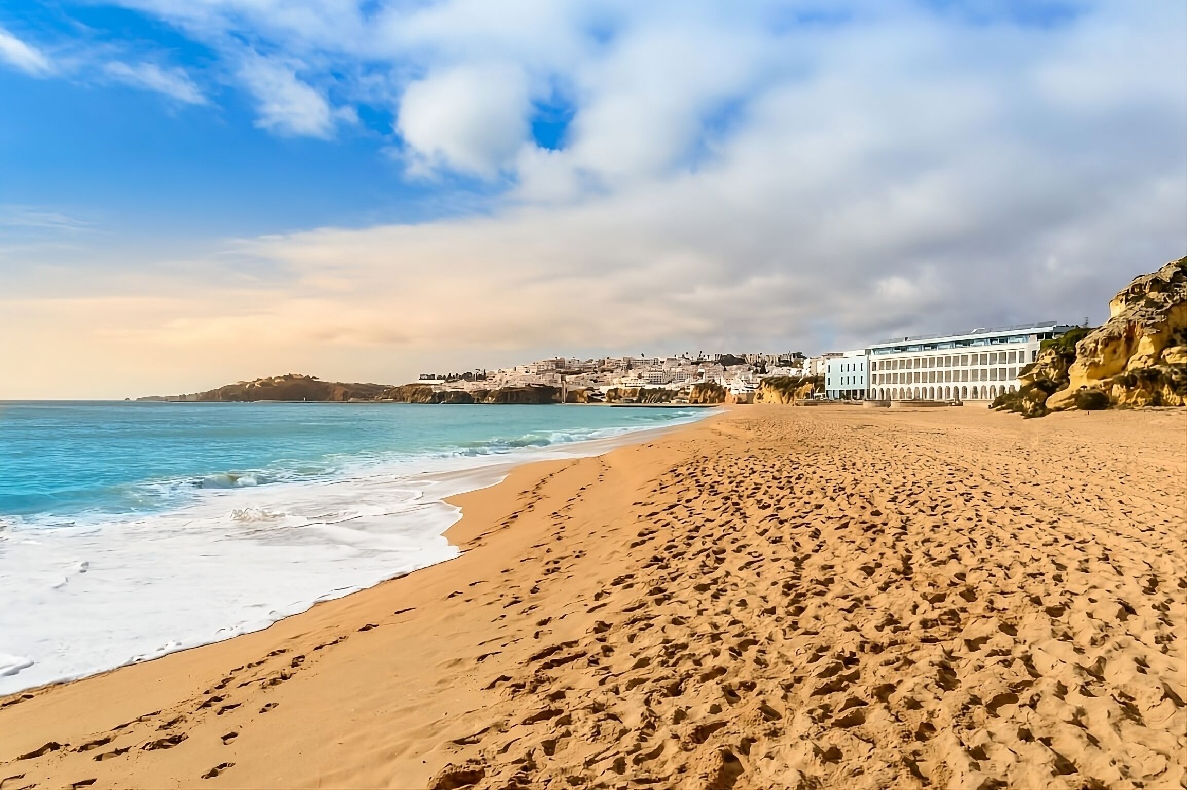 A beach with footprints leading to the water, a rocky cliff, and a building with multiple windows.