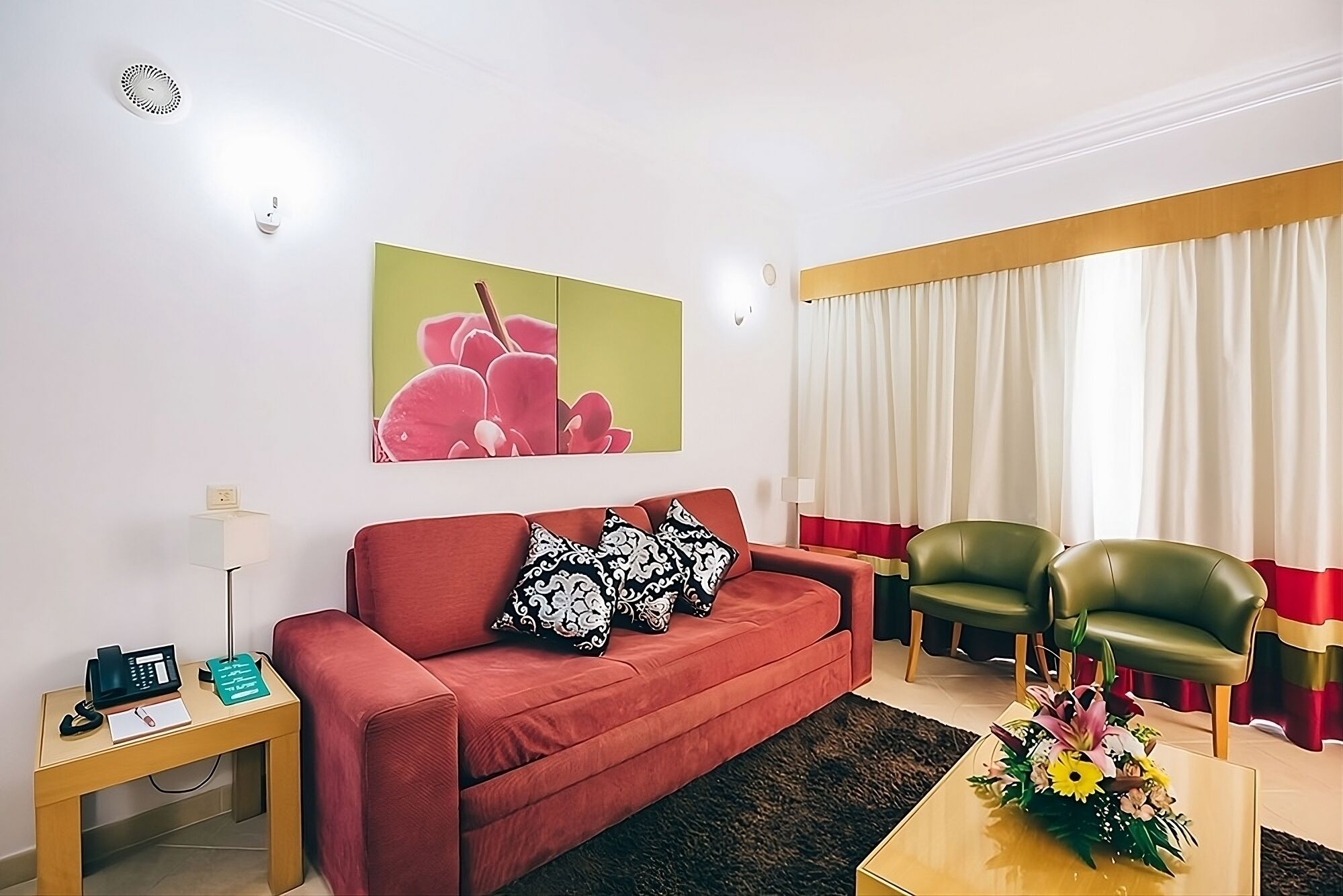 A living room with a red sofa, two green armchairs, a coffee table with a floral arrangement, a painting of flowers on the wall, and a window with curtains.