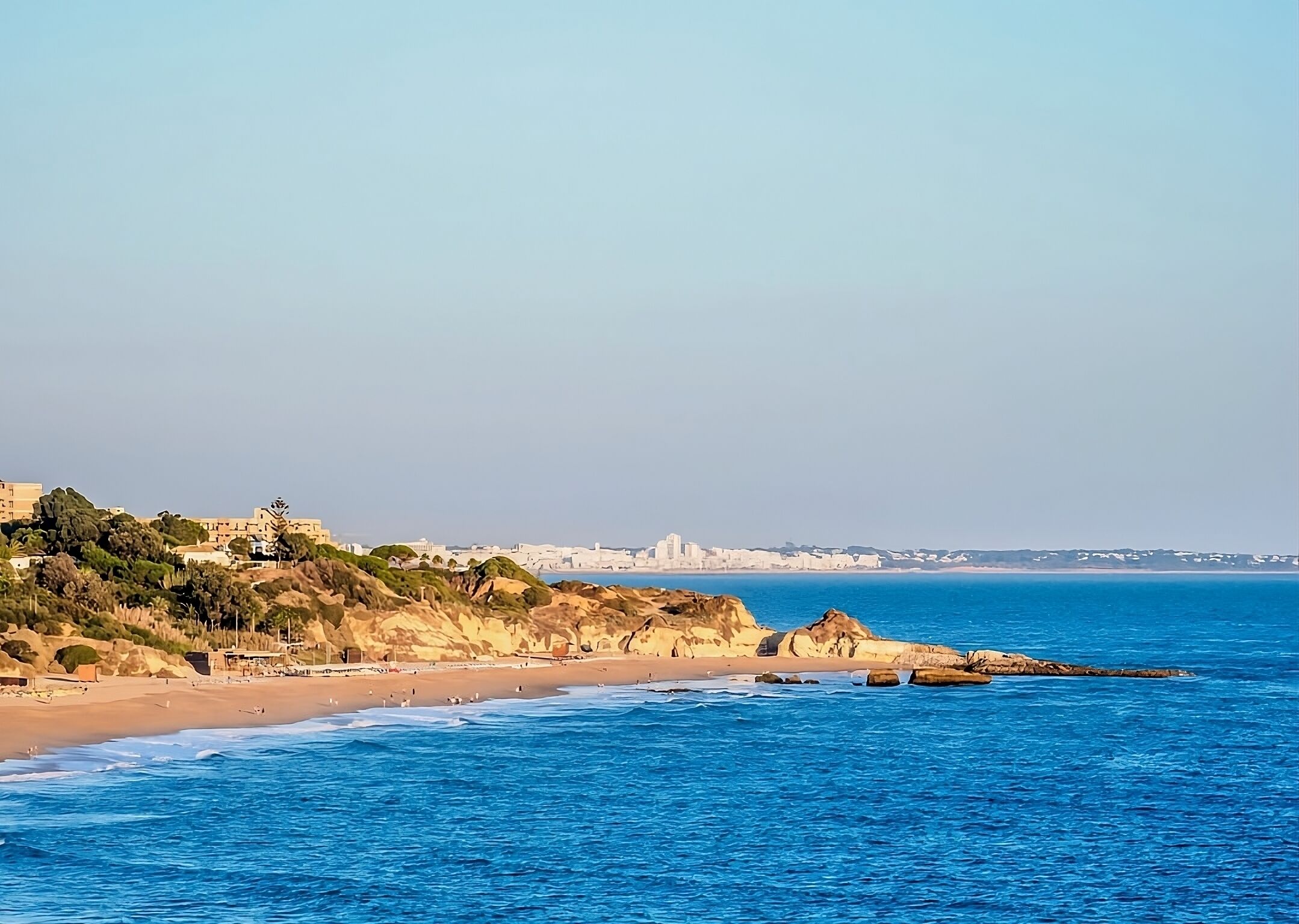 Vista costera con una playa arenosa, afloramientos rocosos y un horizonte urbano distante.