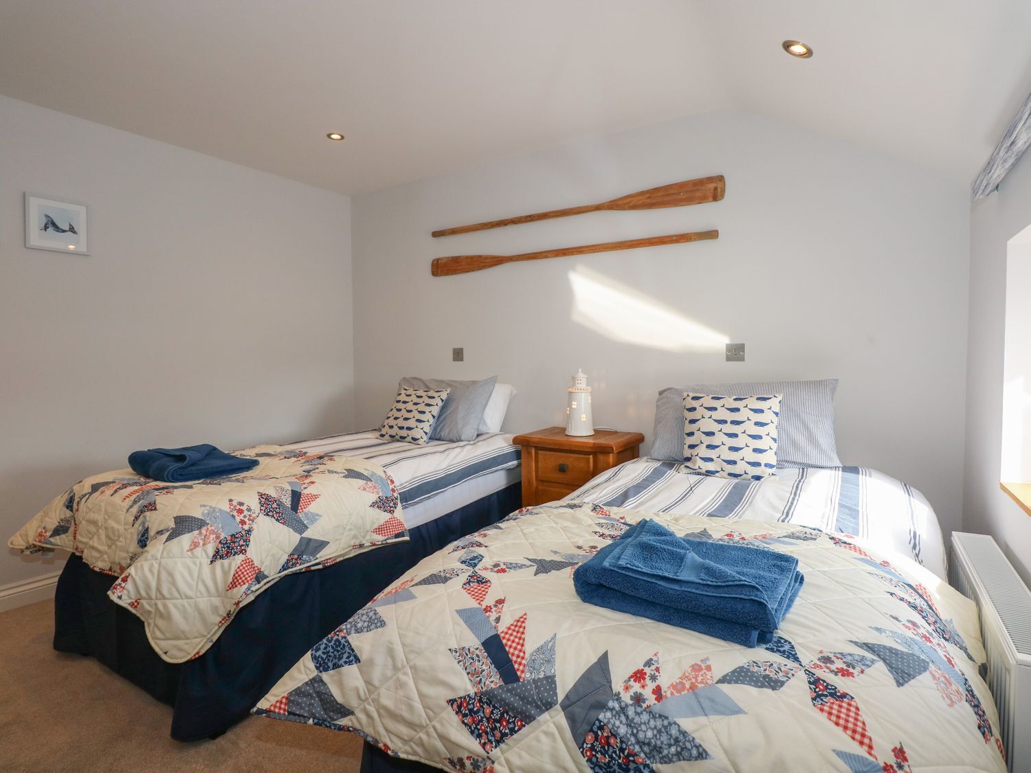 Une chambre avec deux lits, chacun recouvert d’une courtepointe matelassée et agrémenté d’oreillers, une table de chevet avec une lampe et une étagère en bois fixée au mur.