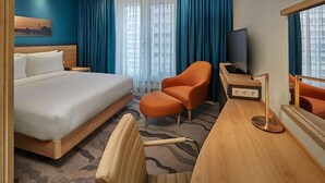 Ein Hotelzimmer mit einem großen Bett, einem Schreibtisch mit Fernseher, einem orangen Sessel und Blick auf Gebäude durch das Fenster.