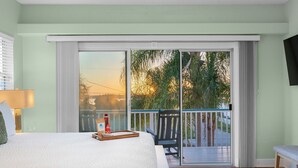 Una camera da letto con un letto grande, un comodino, una lampada, un vassoio con una bottiglia e una sedia vicino a una porta vetrata scorrevole che dà su un balcone con vista su palme e un tramonto.