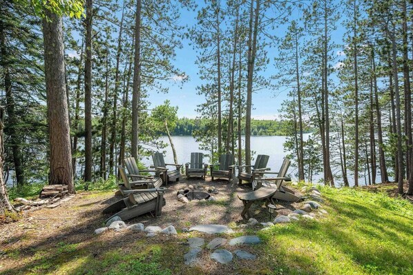 A lakeside campsite with Adirondack chairs and a fire pit surrounded by trees.