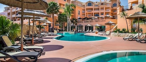 A resort pool area with lounge chairs, umbrellas, and a building with balconies.