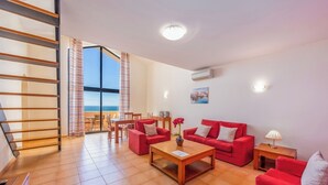A spacious living area with a red sofa set, a wooden coffee table, and a dining table with chairs. There is a large window with curtains, a staircase, and an air conditioning unit on the wall.