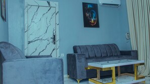 A living room with a grey sofa, a white coffee table with gold legs, a marble-patterned door, and a wall-mounted air conditioner. - Glossy Luxury Hotel (Akure)