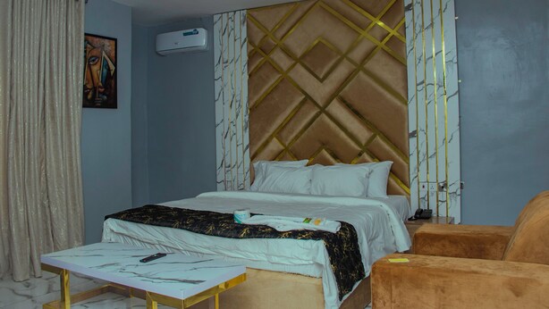 A hotel room with a bed, a bedside table, a chair, and a wall-mounted air conditioner. - Glossy Luxury Hotel (Akure)
