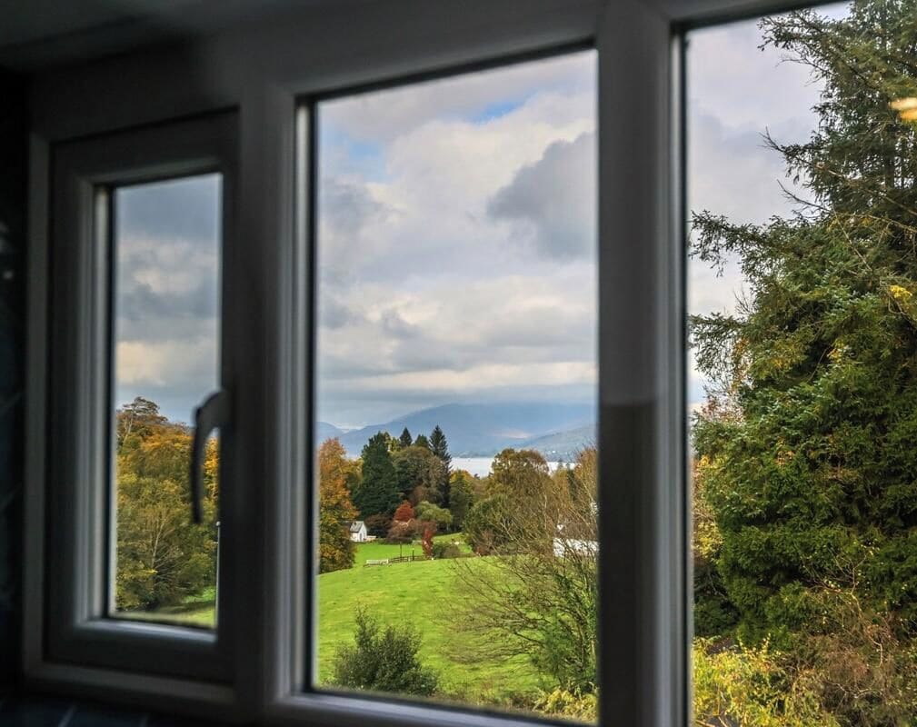 Une vue à travers une fenêtre, donnant sur un paysage composé d’arbres, d’un champ herbeux et d’une étendue d’eau.