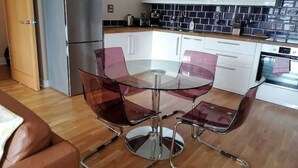 A modern kitchen with white cabinets, a stainless steel refrigerator, a glass dining table with four chairs, and a wooden floor. - Modern City-Centre Apartment with Stunning Views & Free Parking (Cardiff)