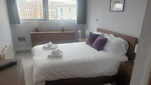 A neatly made bed with white linens and two purple pillows, a bedside table with a lamp, a window with curtains, and a small potted plant on a wooden bench. - Modern City-Centre Apartment with Stunning Views & Free Parking (Cardiff)