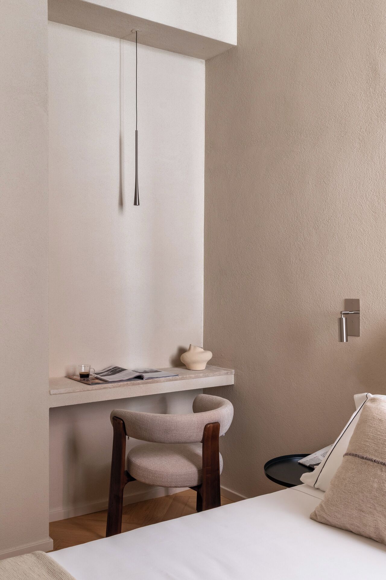A minimalist room with a bed, a desk with a chair, a hanging light, and a wall-mounted shelf.