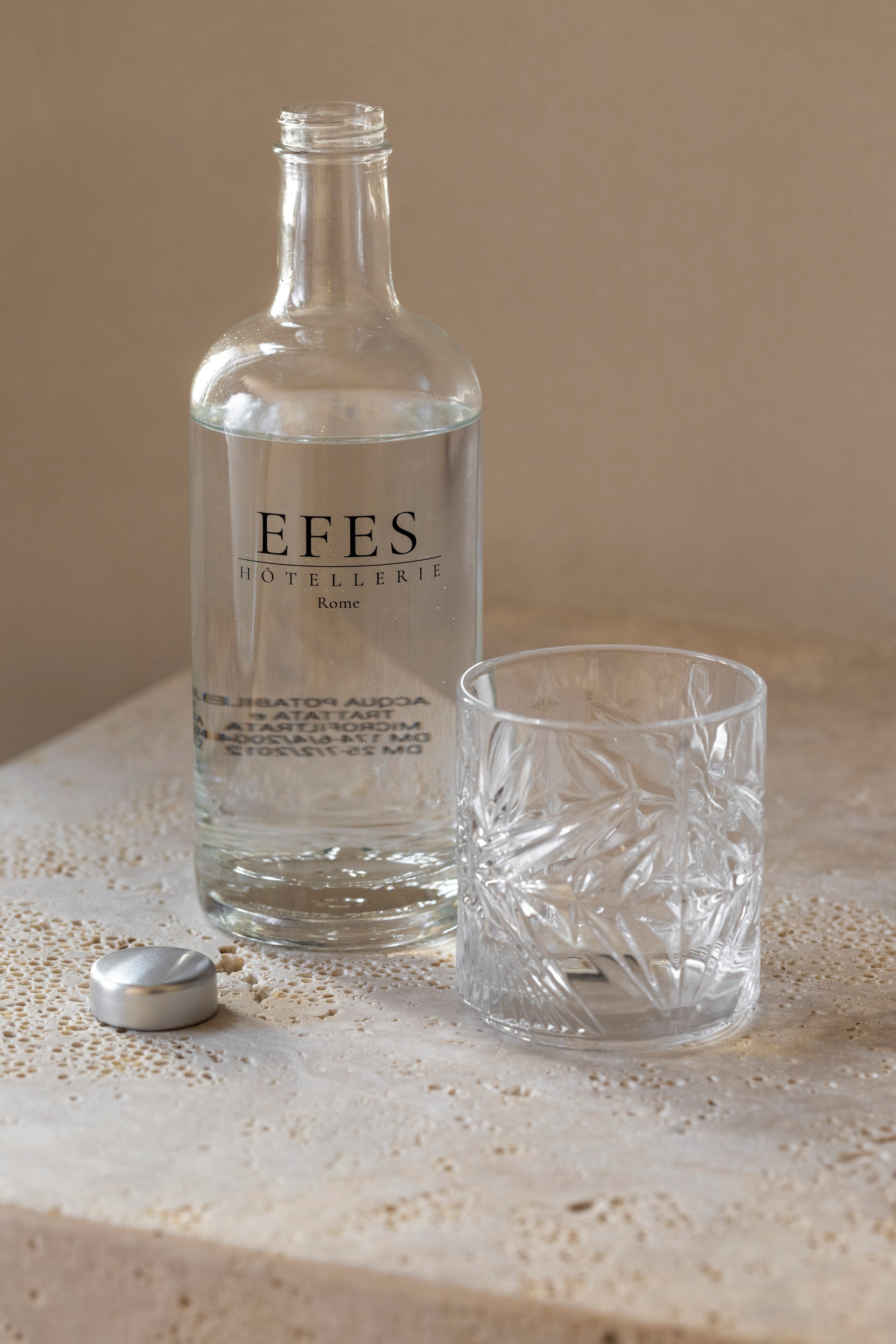 A clear glass bottle labeled 'EFES HÔTELERIE Rome' and a matching glass on a textured surface.