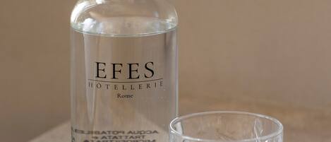 Una botella de vidrio transparente etiquetada como 'EFES HÔTELERIE Rome' y un vaso a juego sobre una superficie texturizada.