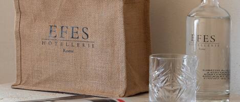 Un sac en toile de jute sur lequel est imprimé le texte « EFES HOTELERIE Rome », une bouteille en verre transparent avec un bouchon argenté et un verre posé sur une table, à côté d’un magazine.