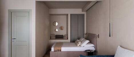 A modern hotel room with a large bed, a smaller bed, a seating area with cushions, a round mirror, and a bedside table with a lamp.
