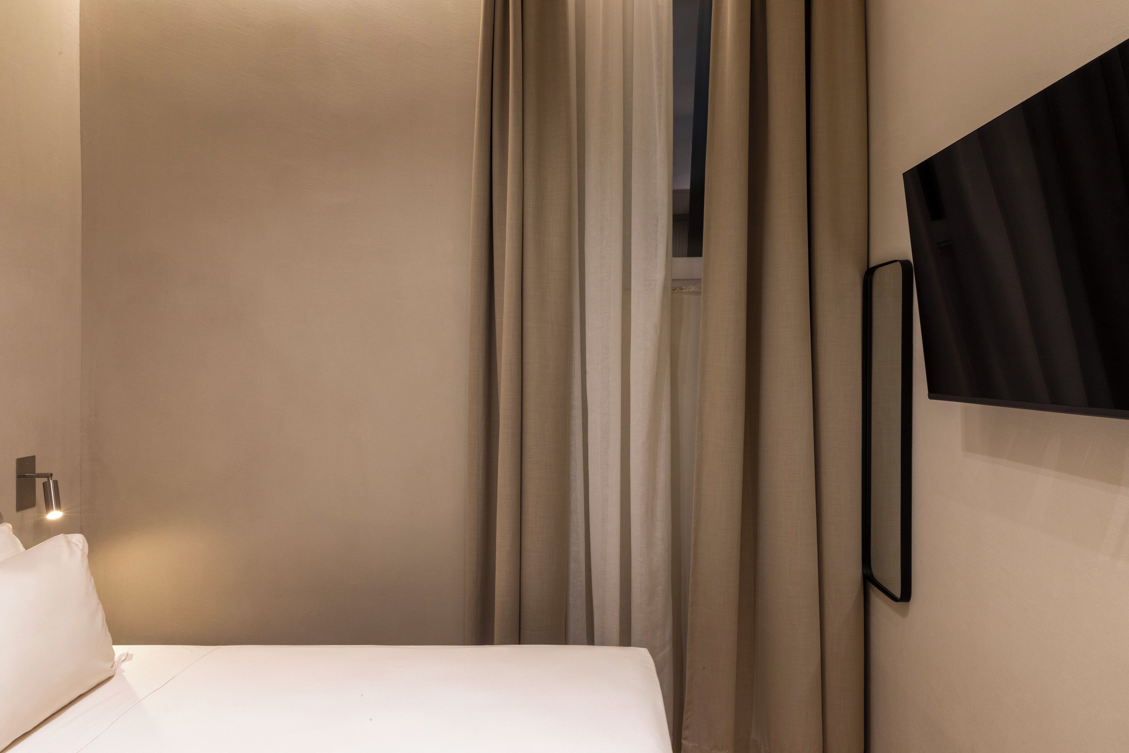 A hotel room with a bed, curtains, a wall-mounted TV, and a bedside lamp.