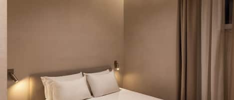 A neatly made bed with white linens in a room with beige walls and a window with curtains.