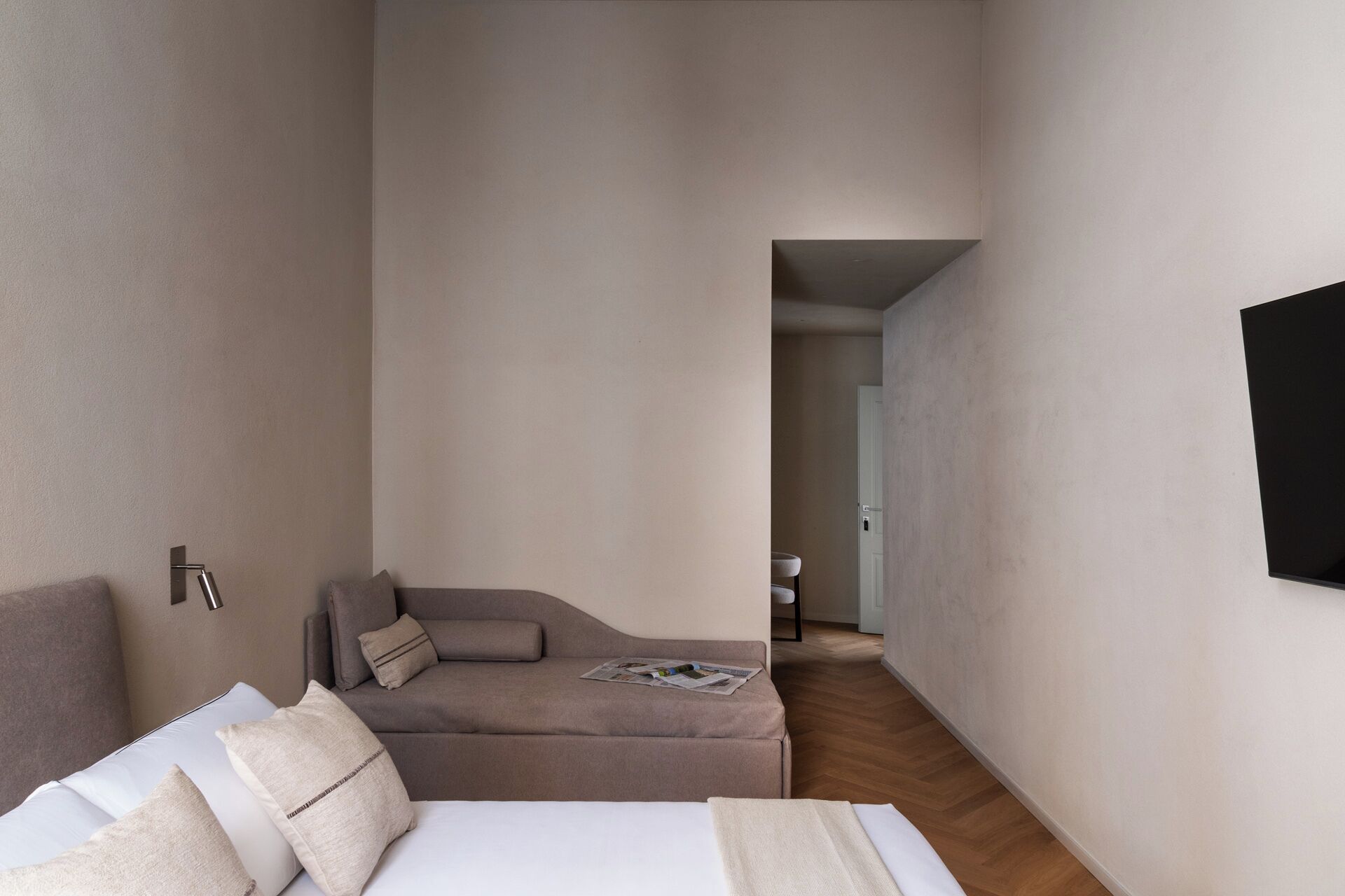 A narrow hotel room with a bed, a sofa, and a TV mounted on the wall.