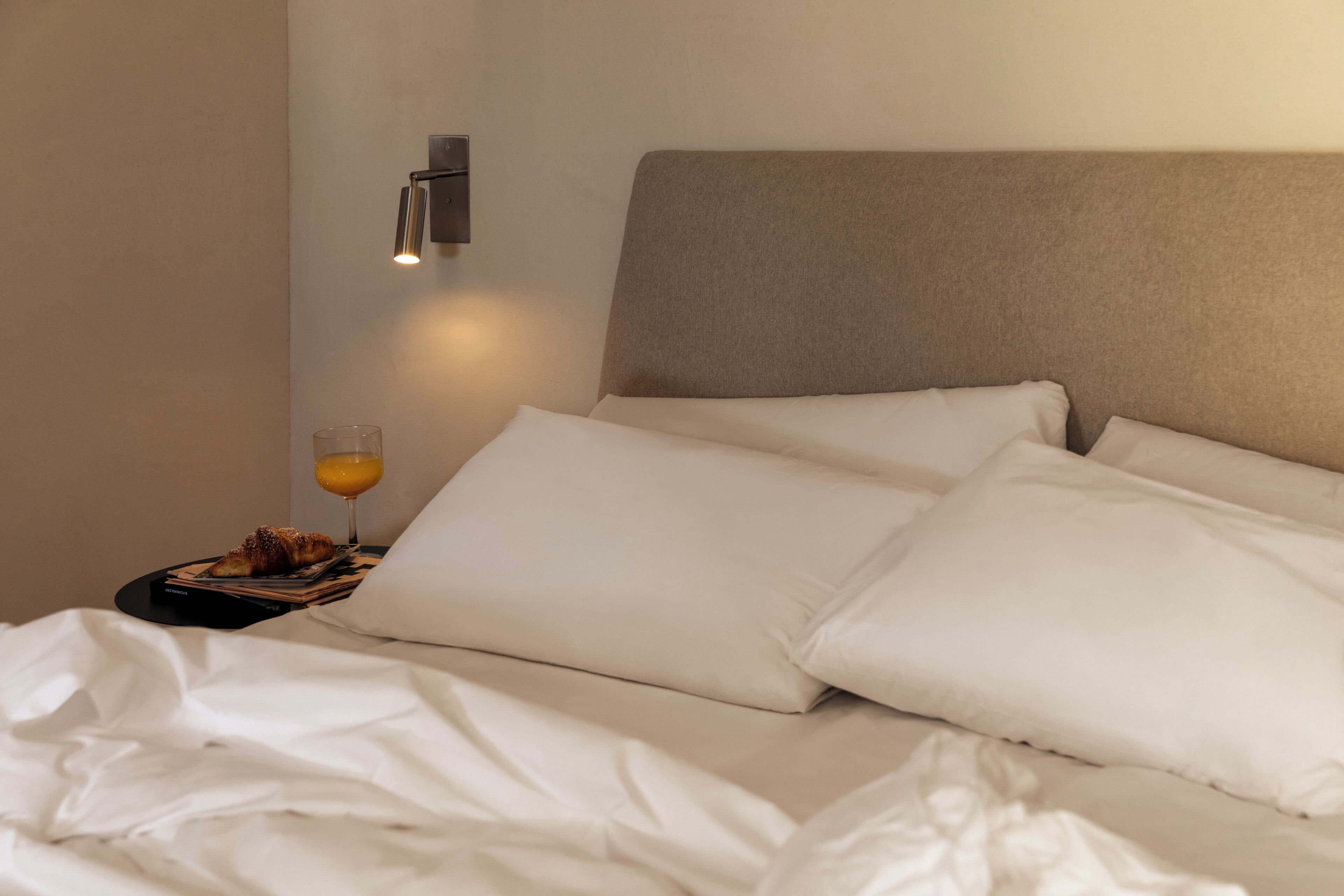 A neatly made bed with white linens, a glass of orange juice, and a book on a bedside table.