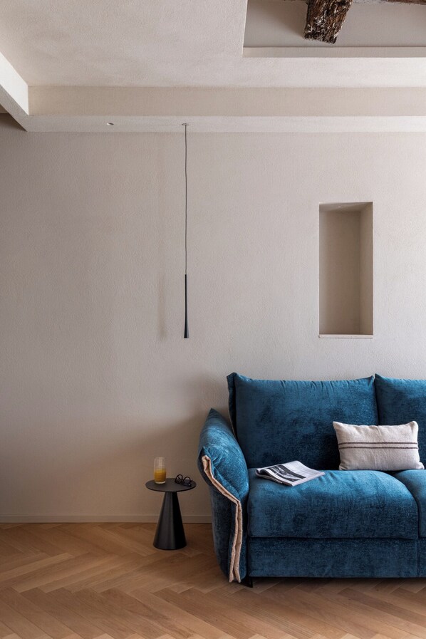 Un salon moderne avec un canapé bleu, une petite table ronde sur laquelle se trouve un verre de jus d’orange, et un luminaire simple.