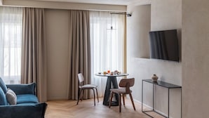 Une chambre d’hôtel avec un canapé, une table à manger avec des chaises, un téléviseur fixé au mur et une grande fenêtre avec des rideaux.