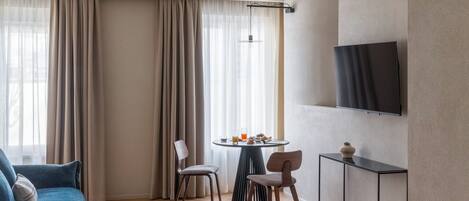 A hotel room with a sofa, a dining table with chairs, a TV mounted on the wall, and a large window with curtains.
