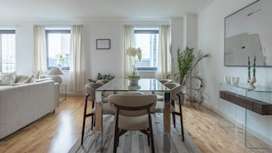 A modern living room with a glass dining table, beige sofa, and large windows with curtains. - High -Rise waterfront Apartment (London)