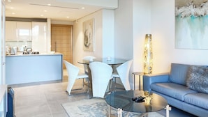 A modern living room with a blue sofa, a glass coffee table, and a dining area with white chairs. - The Marina V&A Waterfront by Smart City Stays (Cape Town)