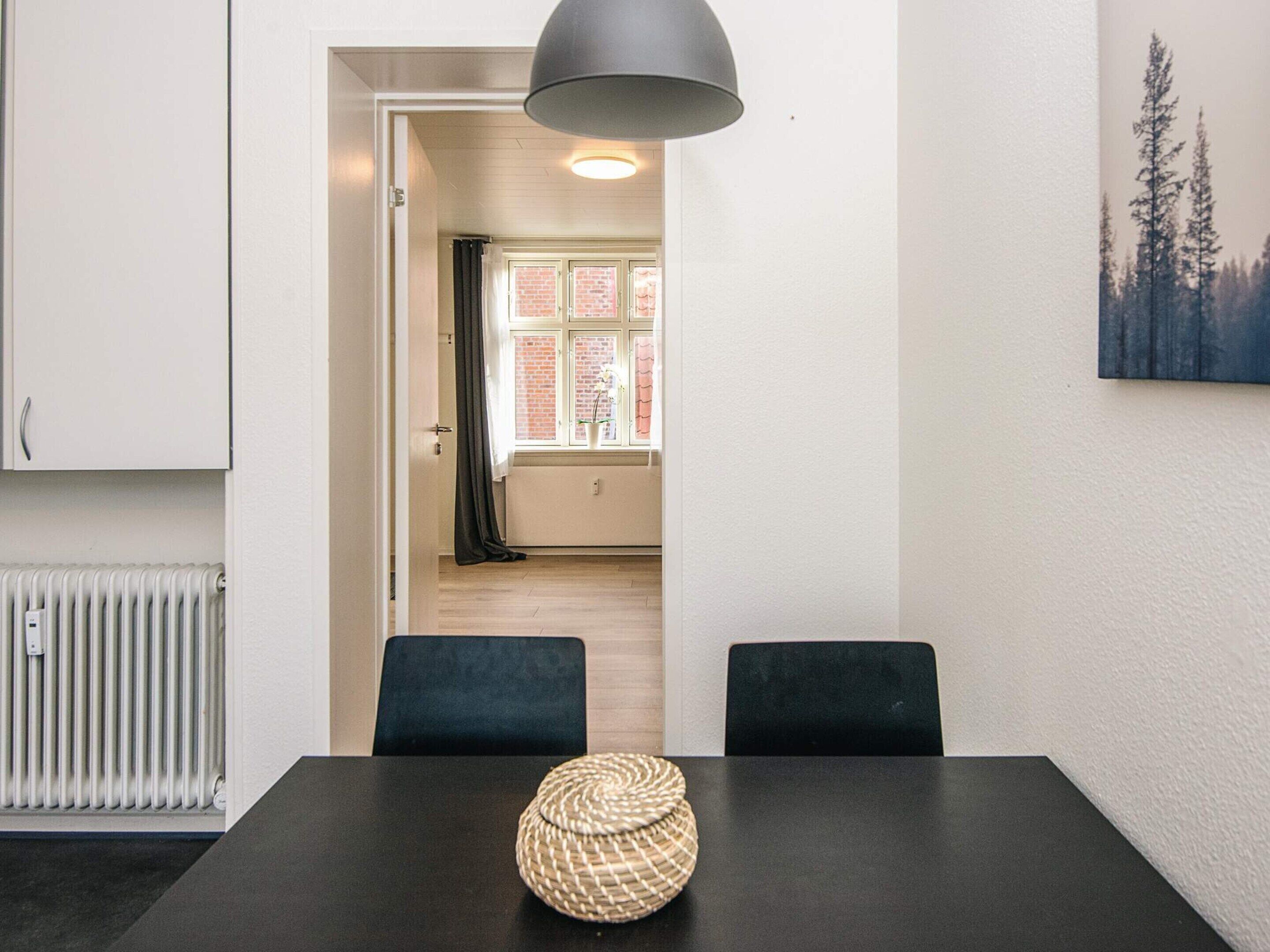 A dining area with a black table, two black chairs, a woven centerpiece, a radiator, and a window with a view of trees.