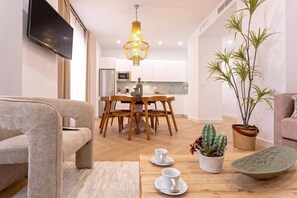 A modern living room with a sofa, a dining area with a wooden table and chairs, a kitchen in the background, and a large plant. - Exclusive Apartment in City Center. Capiteles (Granada)