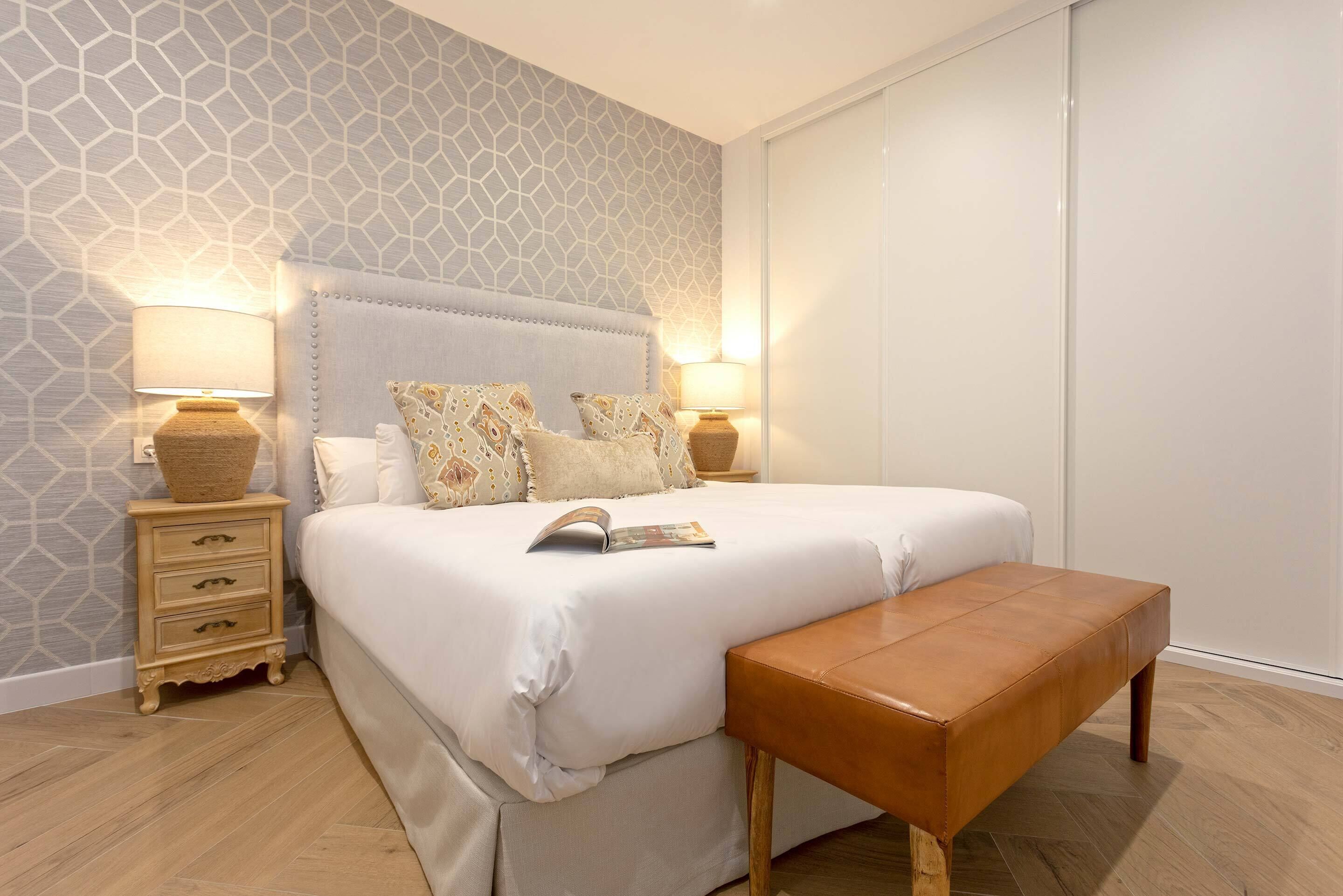 A neatly arranged bedroom with a bed, bedside lamps, a wooden bench, and a patterned wall.