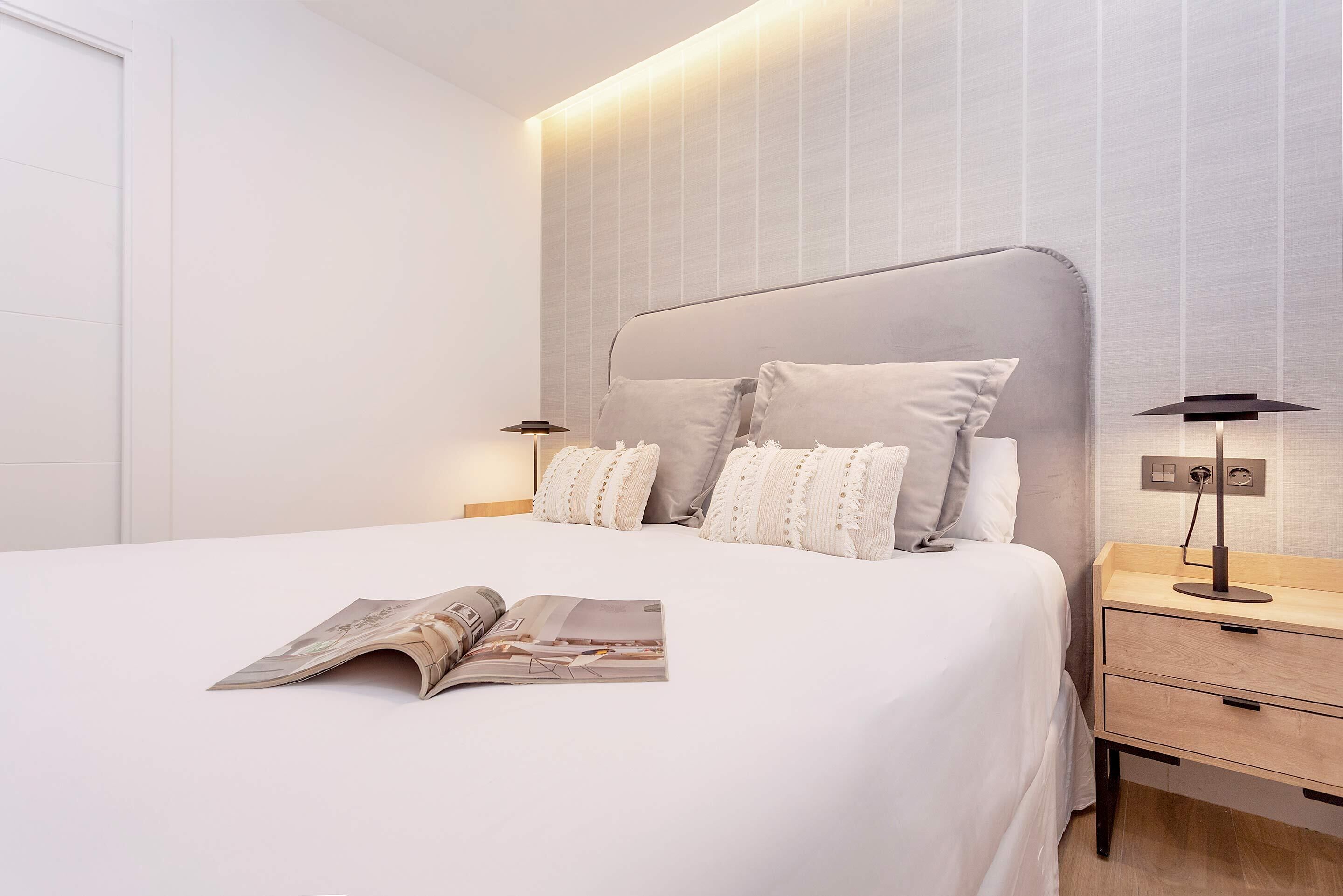 A neatly made bed with a magazine on it, a bedside table with a lamp, and a wall with vertical lines.