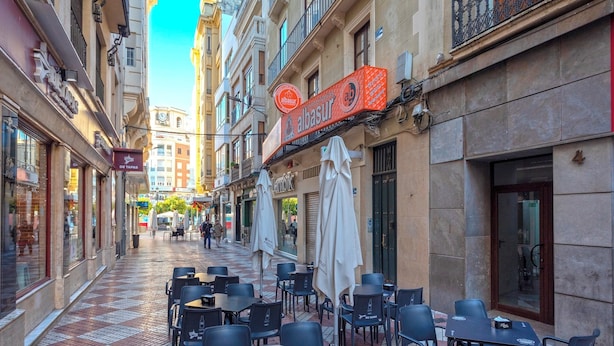 Una calle estrecha con mesas al aire libre, una torre del reloj y un edificio con un letrero que dice "albasur".