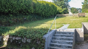 Un escalier en béton mène vers le haut d’une pente herbeuse, avec une main courante en métal. L’ensemble est entouré d’un feuillage verdoyant et dense, et un grillage en bois se trouve à l’arrière-plan.
