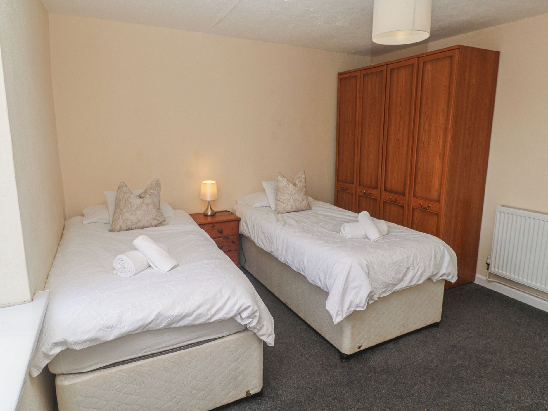 Une chambre avec deux lits, une armoire en bois, une table de chevet avec une lampe et un radiateur.