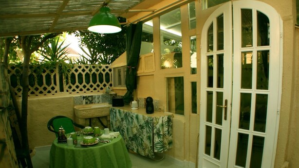 A cozy patio area with a table set for dining, a hanging green lamp, and a door leading outside. - GREEN ROOM OF ARTISTS (Vada)
