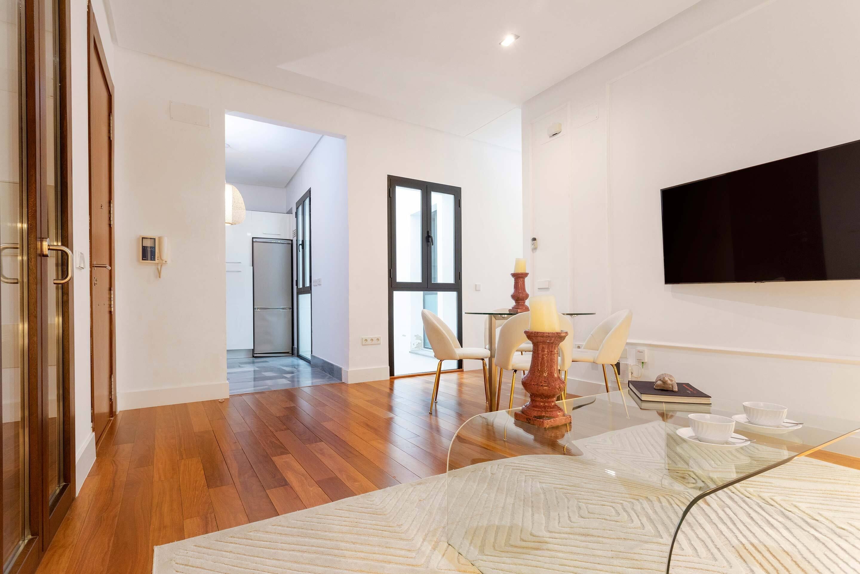 A modern living room with a glass coffee table, a flat-screen TV mounted on the wall, and a dining area with a table and chairs.