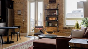 A living room with a sofa, a coffee table, a dining table with chairs, a bookshelf, and a view through a large window. - The Urban Alchemist Loft (Benton Harbor)