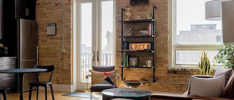 A living room with a sofa, a coffee table, a dining table with chairs, a bookshelf, and a view through a large window.