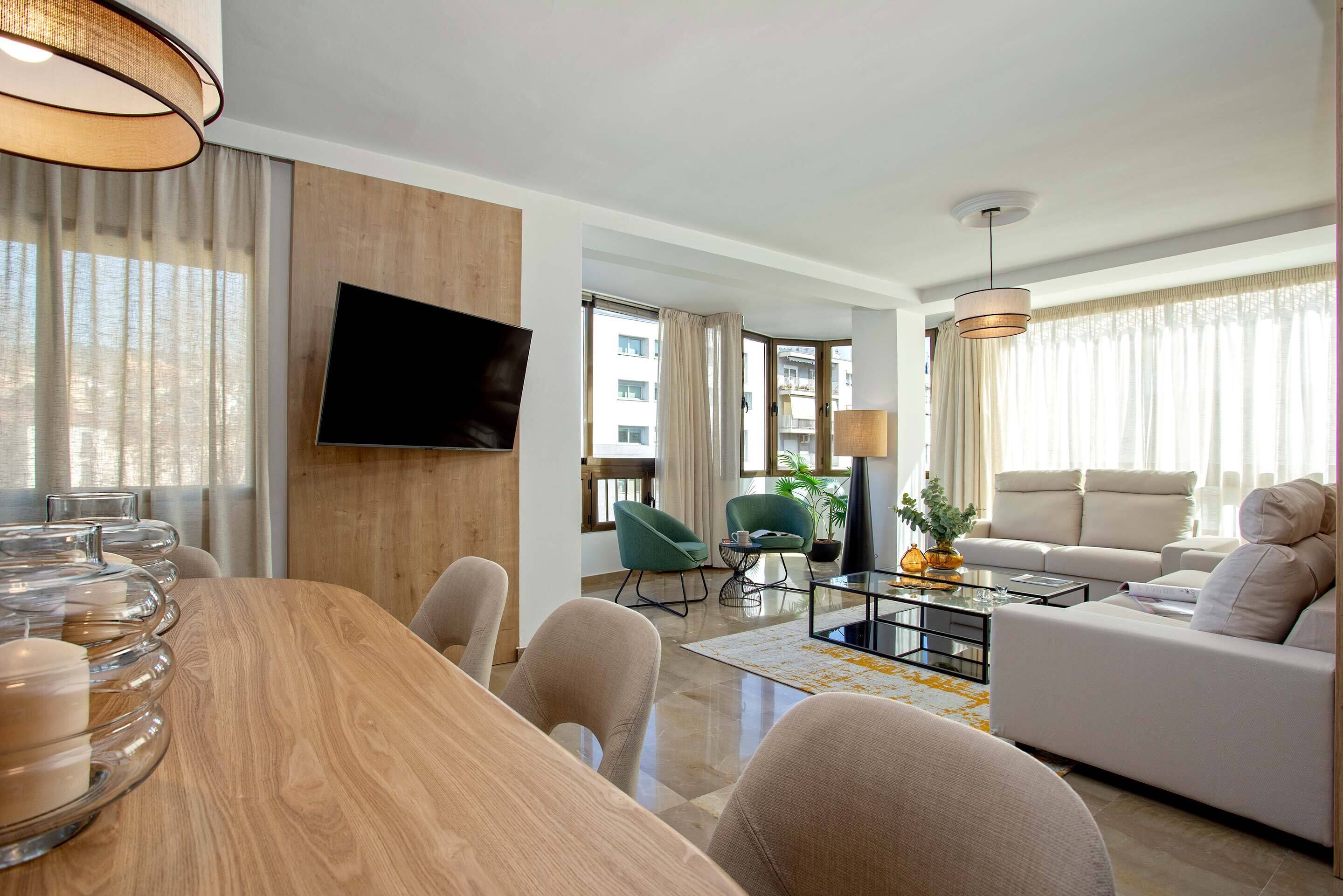 A modern living room with a wooden dining table, a sofa set, and a flat-screen TV mounted on the wall.
