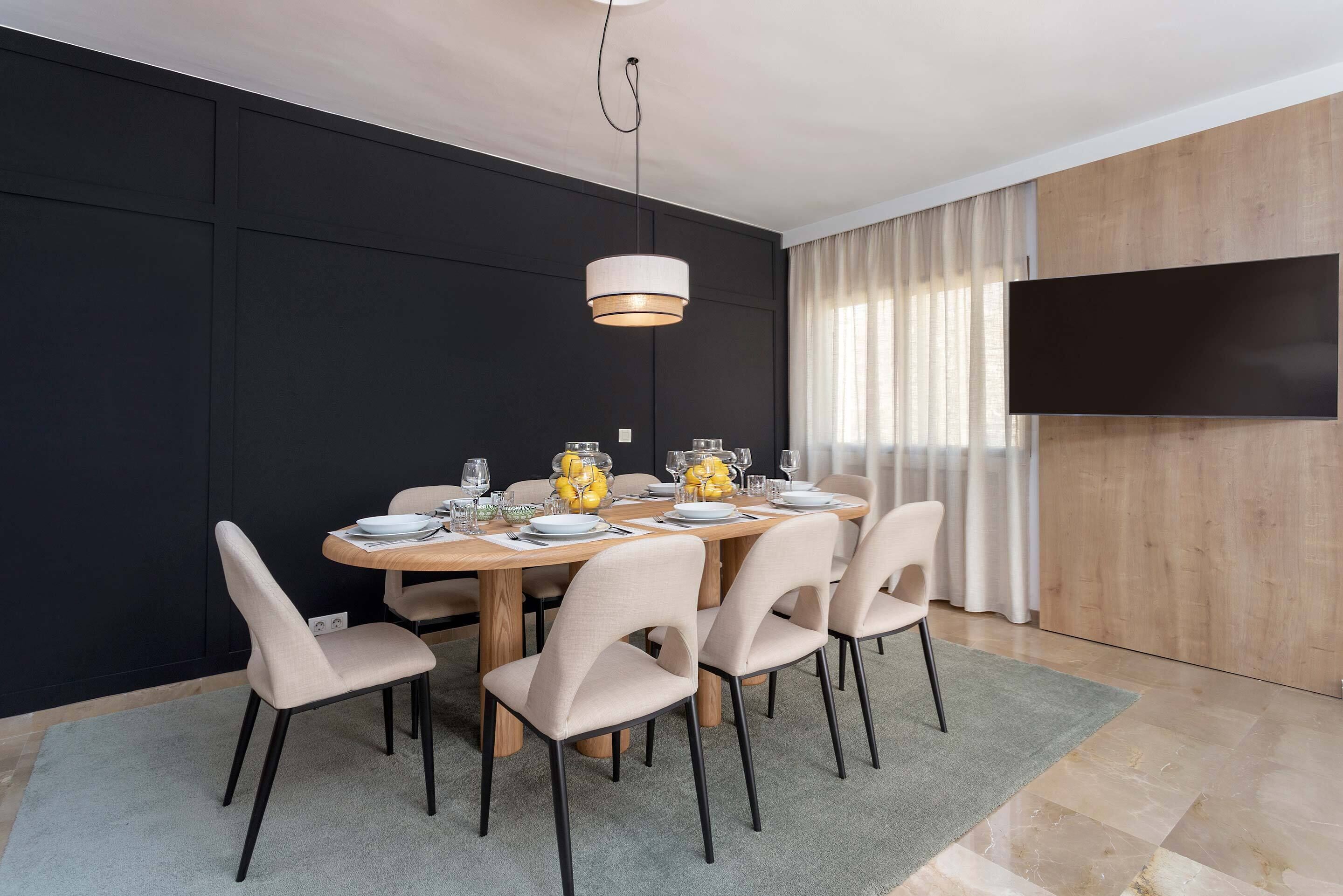 Un comedor con una mesa de madera dispuesta para seis personas, rodeada de sillas color beige, una lámpara colgante moderna y un televisor de pantalla plana grande montado en la pared.