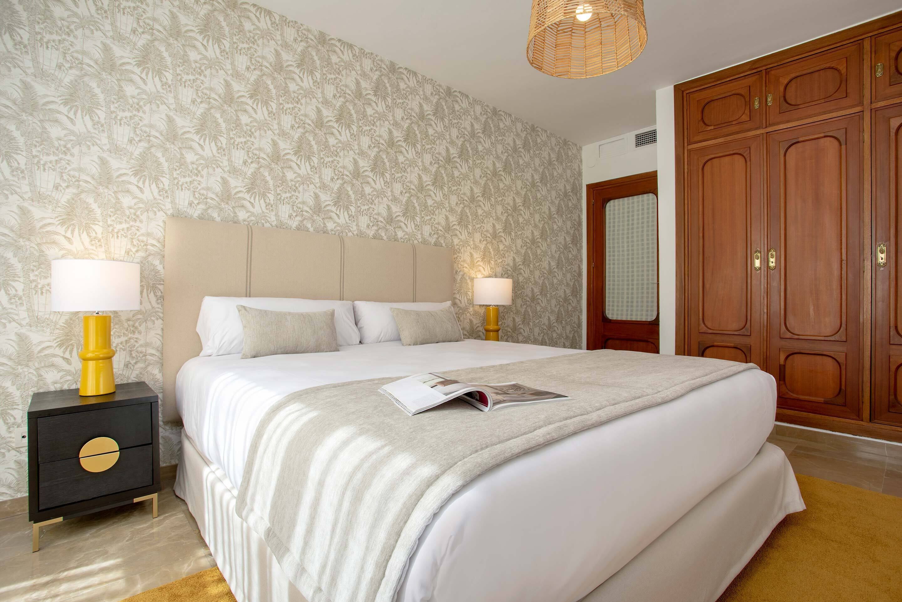 A neatly made bed with a white comforter and pillows, a bedside table with a lamp, and a large wooden wardrobe.