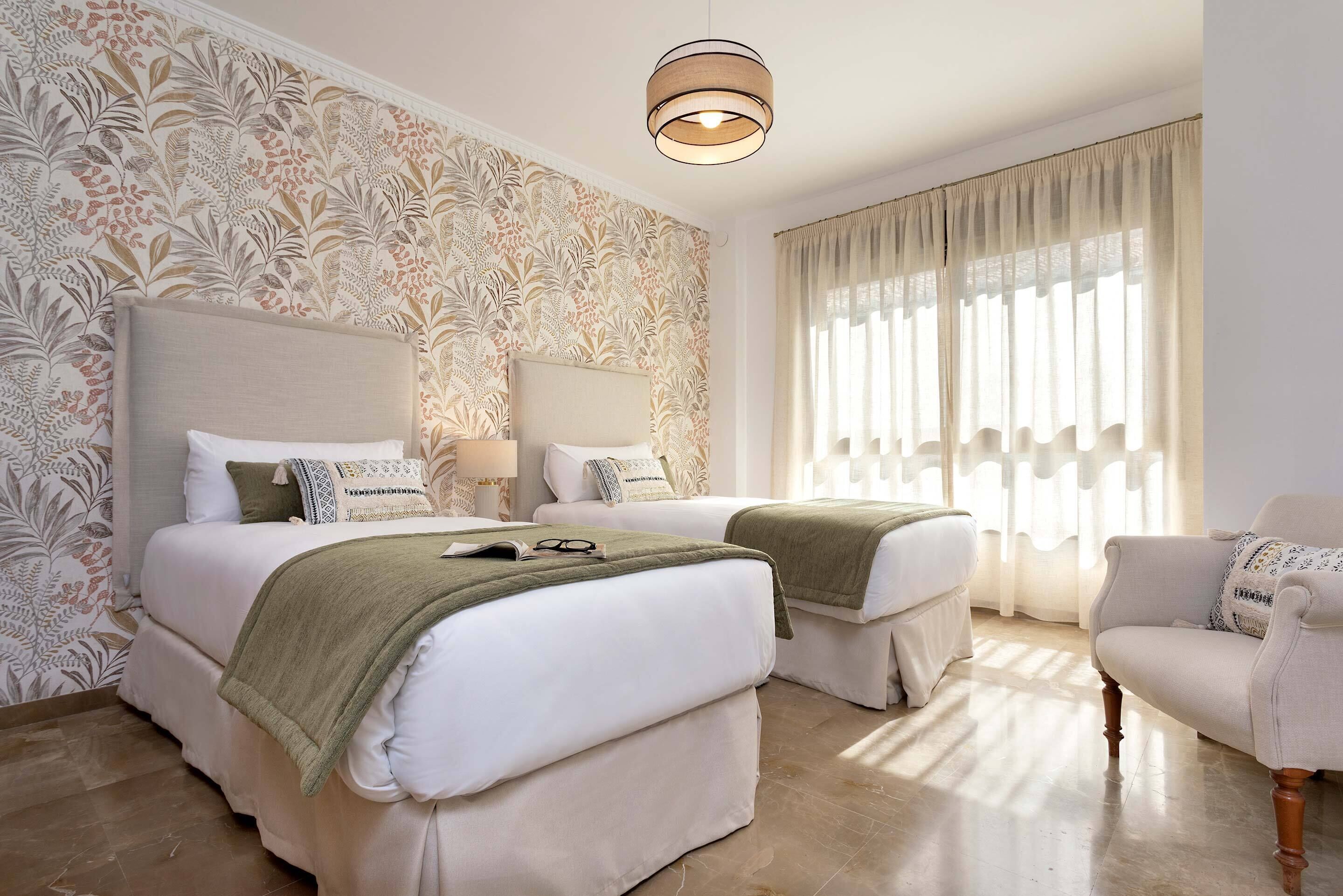 A hotel room with two beds, a sofa, a large window with curtains, and a patterned wallpaper.