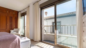 A hotel room with a large sliding glass door to a balcony, a bed with a pink blanket, a white armchair, and a small table with a magazine. - Luxury Apartment in City Center. Puerta Real II (Granada)
