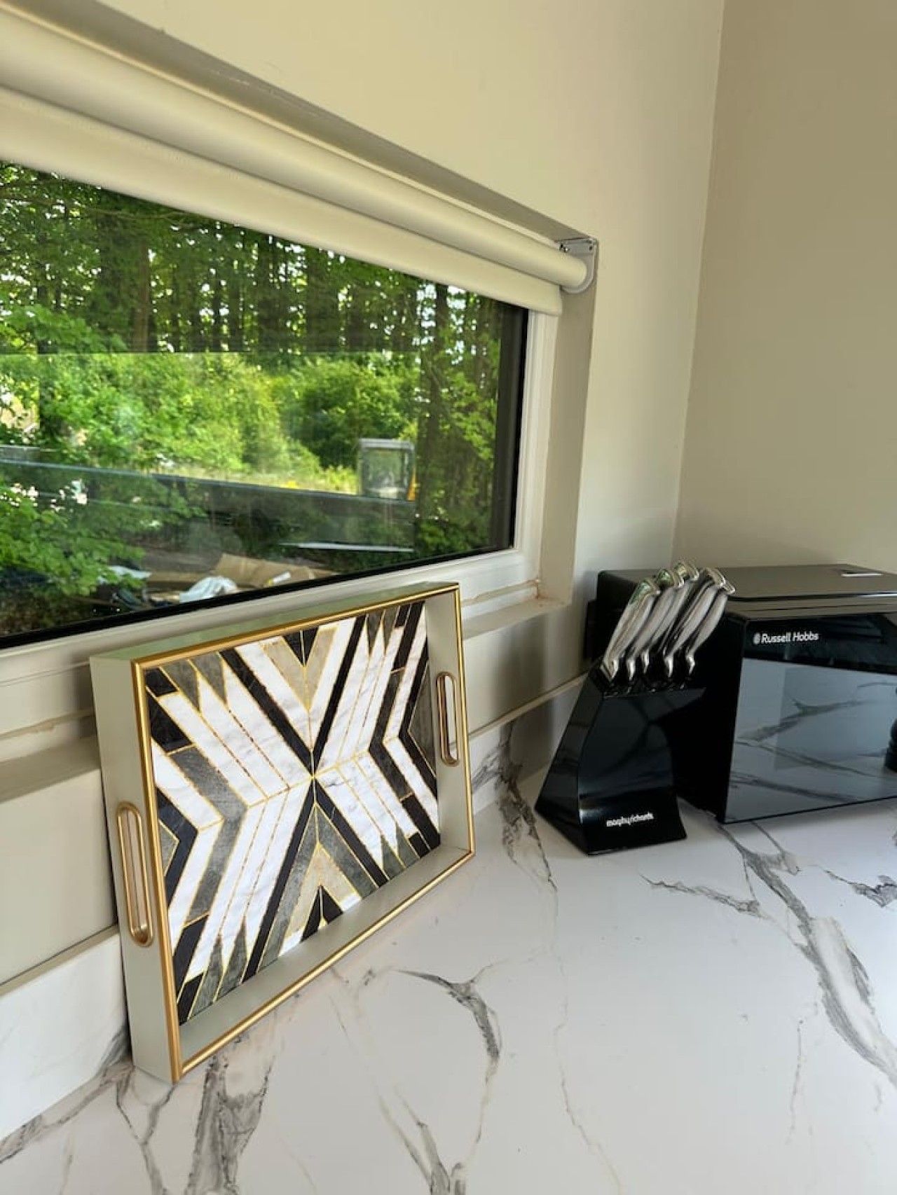 Ventana con vista a follaje, obra de arte geométrica enmarcada y una caja negra con juego de cuchillos.