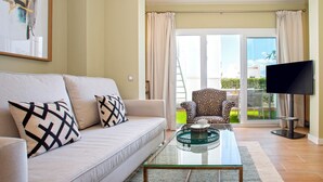 A living room with a sofa, armchairs, a glass coffee table, a flat-screen TV, and a sliding glass door leading to an outdoor area.