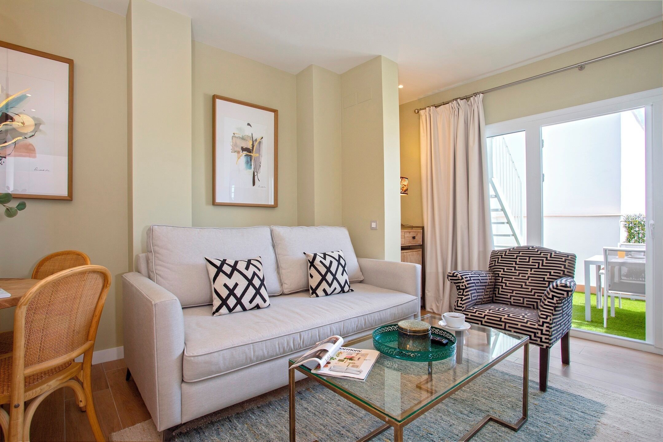 Una sala de estar con un sofá, un sillón estampado, una mesa de centro de vidrio y un comedor con sillas de mimbre.