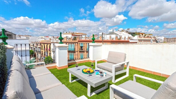 A rooftop terrace with outdoor furniture, a dining table with a fruit bowl and wine glasses, and a view of the cityscape.