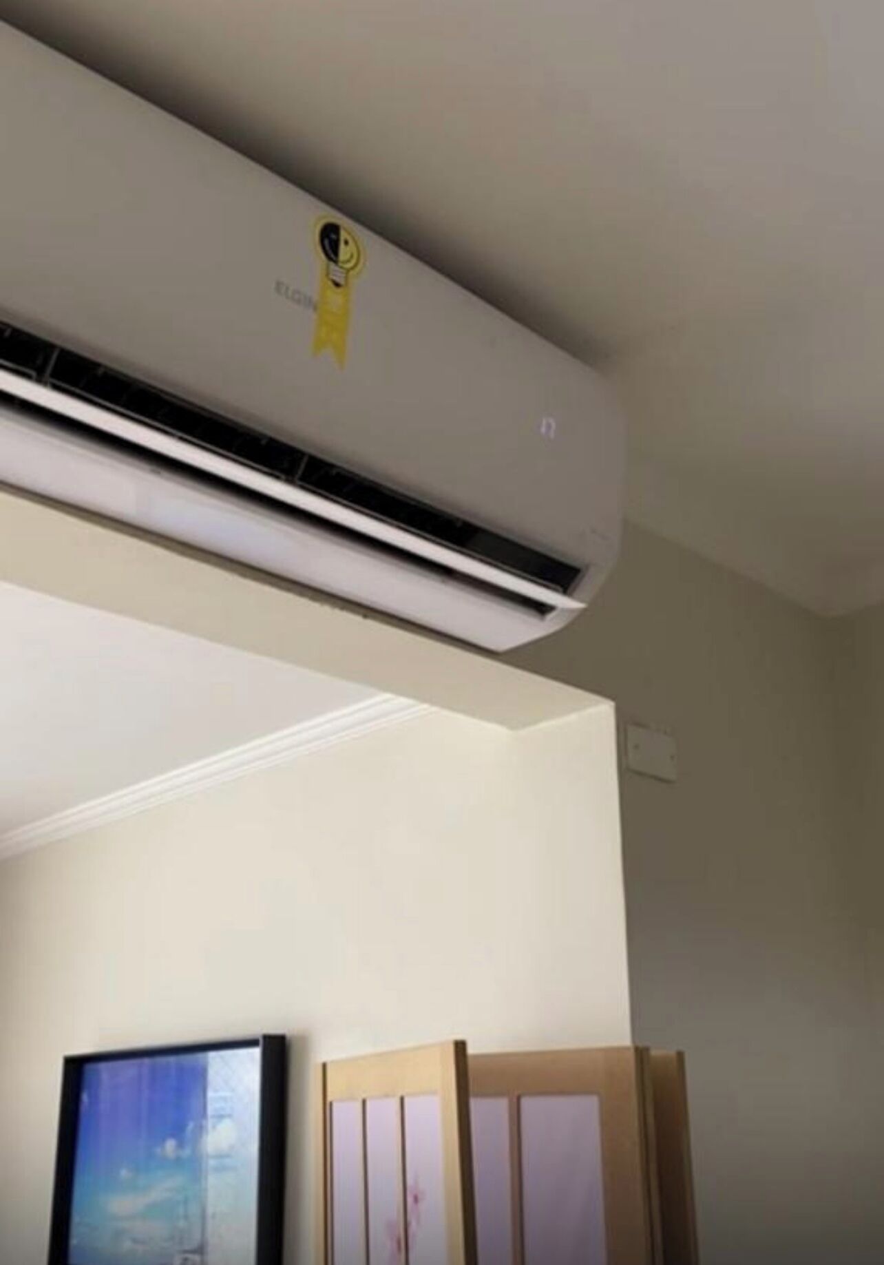 A room with a ceiling-mounted air conditioner, a framed picture on the wall, and a wooden cabinet.