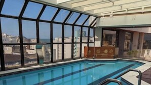 An indoor swimming pool with clear glass walls, a tiled floor, and a view of the cityscape. - Flat Golden Beach Pitangueiras with Sea View (Guarujá)